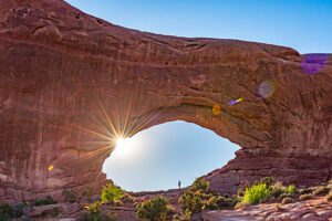 Person standing in a canyon with a large natural arch in the distance under a clear blue sky