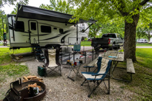 RV campsite with camping chairs, picnic table, fire ring, and outdoor gear set up beside a travel trailer, showcasing a comfortable camping experience for RV travelers.