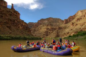 Group river rafting adventure with multiple inflatable rafts on a calm river surrounded by towering canyon cliffs and scenic desert landscape.