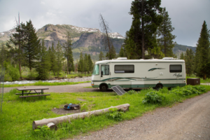 RV camper parked at a scenic campground in Yellowstone National Park with a mountain backdrop, evergreen forest, flowing river, and peaceful campsite surrounded by nature.