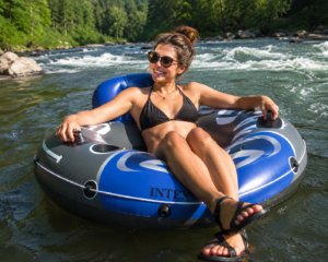 Floating Docks & Water Relaxation Gear: 30 Best Floating Platforms, River Loungers & Lake Essentials 5 Person relaxing in an inflatable river tube while floating down a scenic river surrounded by forest, rocks, and gentle flowing water on a sunny day.