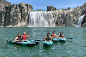 The Ultimate Yellowstone National Park Guide: Valleys, Lakes, Yellowstone River & waterfalls 5 Visitors kayaking on a turquoise lake in front of a spectacular waterfall surrounded by rugged rock cliffs on a sunny day.