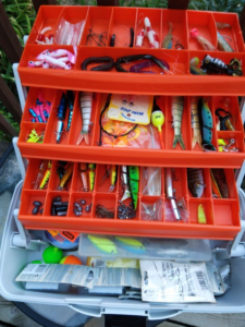 Open fishing tackle box filled with colorful fishing lures, soft plastics, hooks, sinkers, bobbers, swivels, weights, and essential tackle accessories organized in multiple compartments for freshwater and saltwater fishing.