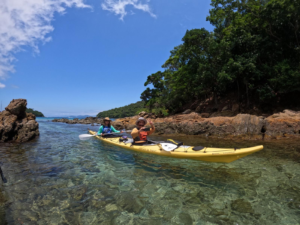 🏝️ Ilha Grande – Beaches, Rainforest Trails, Kayaking & Island Adventures 6 Two kayakers paddling through clear tropical water beside the rocky rainforest coastline of Ilha Grande, Brazil.