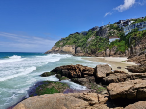 Hidden beach along Rio de Janeiro’s coastline with rocky cliffs, Atlantic waves, sandy shoreline, tropical vegetation and hillside homes beneath a blue sky.