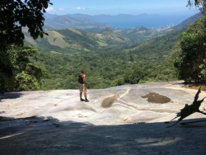 Rio’s Beaches, Coastal Trails & Outdoor Adventures 11 Hiker standing at a rocky viewpoint overlooking forest-covered mountains, valleys, and distant coastline in the Pedra Branca area of Rio de Janeiro.