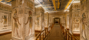 Interior of an ancient Egyptian royal tomb in Luxor with colorful hieroglyphs, painted gods and pharaohs, decorated pillars, and a walkway leading through burial chambers.