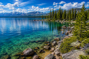 Crystal-clear mountain lake in Yellowstone National Park featuring turquoise water, a rocky shoreline, evergreen forests, and scenic mountain peaks beneath a bright blue sky.