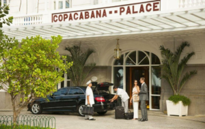 Guests arriving with luggage at the entrance of the Copacabana Palace hotel in Rio de Janeiro, with hotel staff and a vehicle outside.