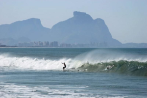 Rio’s Beaches, Coastal Trails & Outdoor Adventures 6 Surfer riding an ocean wave along the Rio de Janeiro coastline with dramatic mountains and the city visible in the distance.