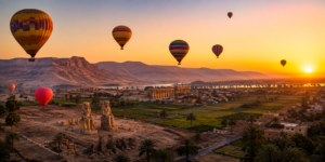 Hot air balloons floating over Luxor at sunrise with ancient Egyptian monuments, green Nile Valley farmland, palm trees, desert mountains, and the Nile in the distance.