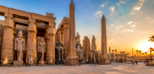 Ancient temples on Luxor’s East Bank in Egypt with towering pharaoh statues, hieroglyph-covered obelisks, massive stone columns, and palm trees at sunset.
