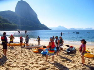 Kayakers preparing for a water adventure on a Rio de Janeiro beach with colorful kayaks, paddles, life jackets, calm Atlantic water and mountains in the background.