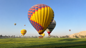 🎈 Hot Air Balloon Rides – Sunrise Over Luxor &the Nile Valley 10 Colorful hot air balloons flying over green fields near Luxor, Egypt, with desert mountains in the distance.
