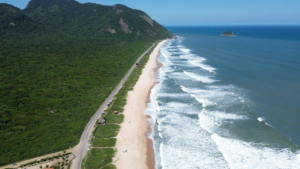 Rio’s Beaches, Coastal Trails & Outdoor Adventures 13 Aerial view of Grumari Beach in Rio de Janeiro with a long sandy shoreline, Atlantic waves, dense tropical vegetation, forest-covered mountains, and an offshore island.