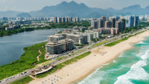Aerial view of beachfront hotels and high-rise buildings along Barra da Tijuca in Rio de Janeiro, with a wide sandy beach, turquoise Atlantic Ocean, lagoon and mountains in the background.