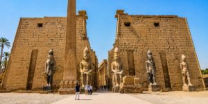 Wide view of Luxor Temple in Egypt featuring the monumental entrance, towering obelisk, colossal statues of Ramesses II, ancient sandstone walls, and visitors exploring the temple complex.