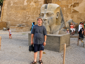 Jeffrey visiting an ancient temple complex in Luxor, Egypt, standing beside a large pharaoh statue with hieroglyph-covered temple walls in the background.