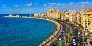 Wide panoramic view of Alexandria, Egypt, showing the Mediterranean Sea, Alexandria Corniche, waterfront buildings, busy coastal roads, and historic harbor scenery.