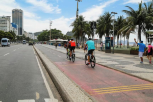 Rio’s Beaches, Coastal Trails & Outdoor Adventures 7 : Cyclists riding along a beachfront bike path in Rio de Janeiro beside palm trees, the promenade, beach, and Atlantic Ocean.