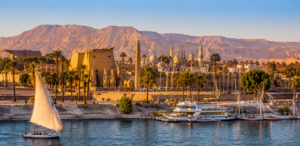 Panoramic view of Luxor, Egypt along the Nile River with ancient temples, palm trees, felucca sailboats, and the desert mountains of the Theban West Bank.