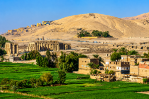 Ancient temple ruins, green farmland, local buildings, palm trees, and desert mountains viewed across Luxor’s West Bank in Egypt.