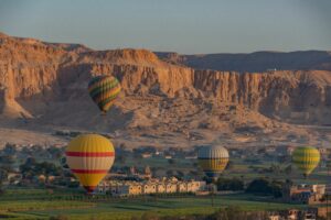 Colorful hot air balloons floating above green farmland and desert mountains on Luxor’s West Bank in Egypt during the early morning.