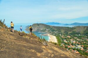 Rio’s Beaches, Coastal Trails & Outdoor Adventures 3 Hikers walking across a rocky coastal trail overlooking a beach, ocean, green hills, and neighborhoods near Rio de Janeiro, Brazil.