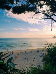 Grumari Beach in Rio de Janeiro viewed through tropical vegetation with a sandy shoreline, Atlantic Ocean, beachgoers and gentle waves beneath a colorful sky.
