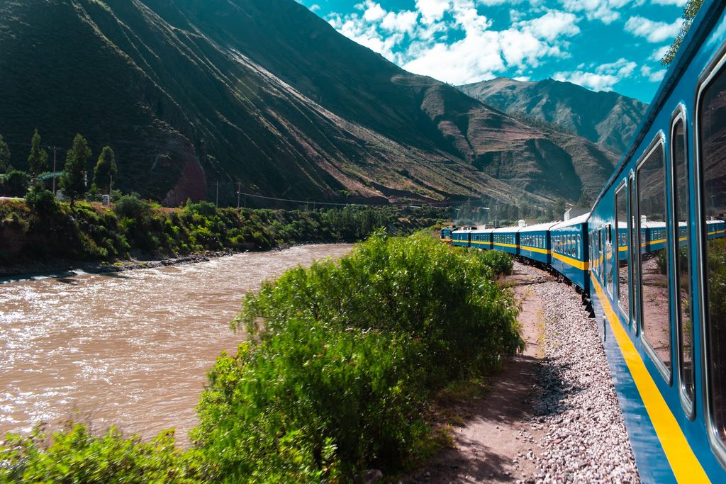 Scenic blue and yellow passenger train winding alongside a river through the mountainous Sacred Valley region of Peru.