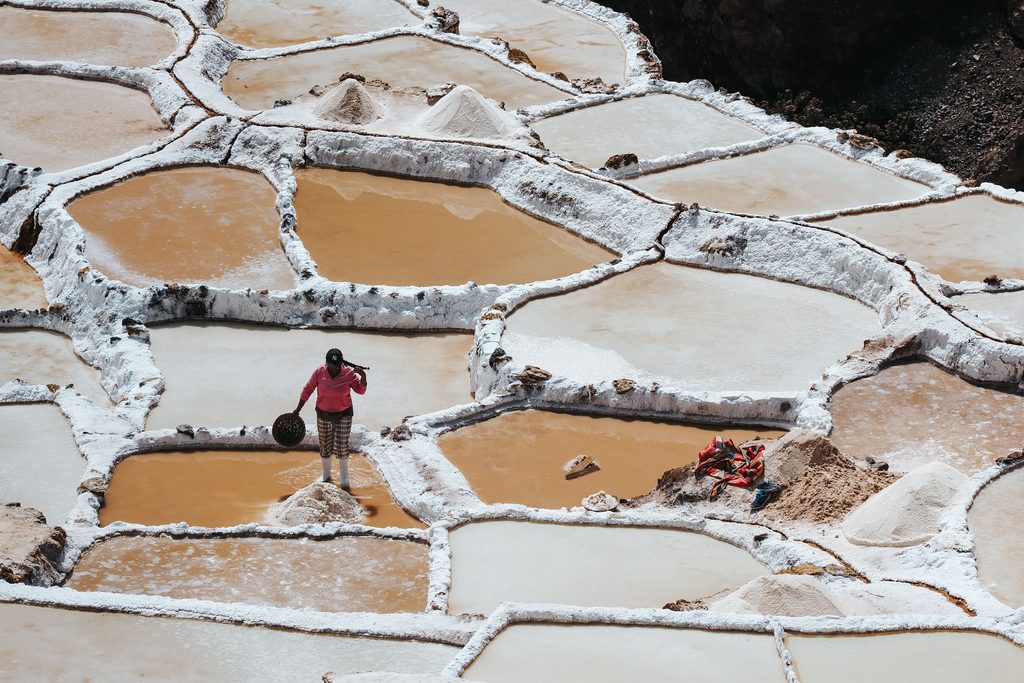 Salt worker standing among traditional salt evaporation pools at the Maras Salt Mines in Peru's Sacred Valley, surrounded by white salt crystals and mineral-rich water.