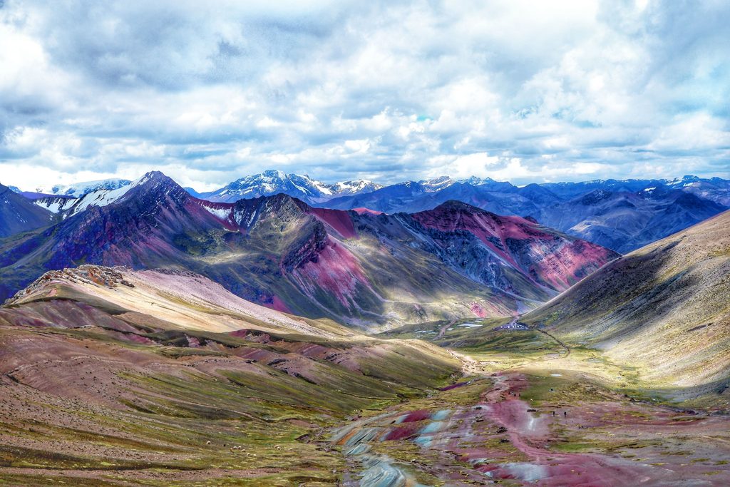 Panoramic view of colorful Andean mountain ridges, snowcapped peaks, and a sweeping high-altitude valley beneath dramatic cloudy skies in Peru.