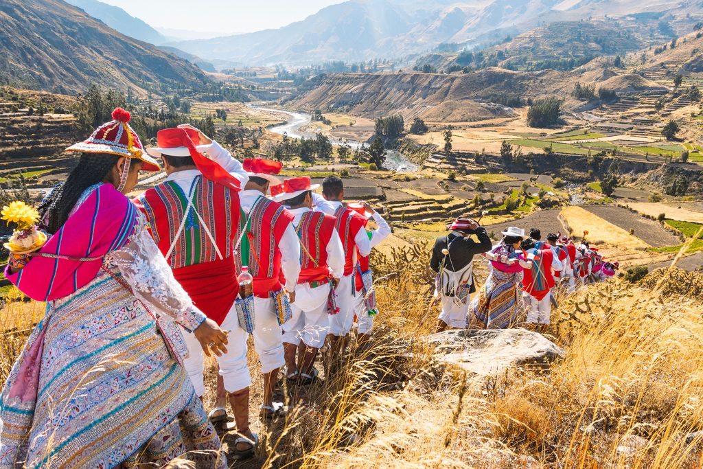 Group of people wearing colorful traditional Andean clothing walking along a hillside trail overlooking a scenic river valley, agricultural terraces, and mountains in Peru.