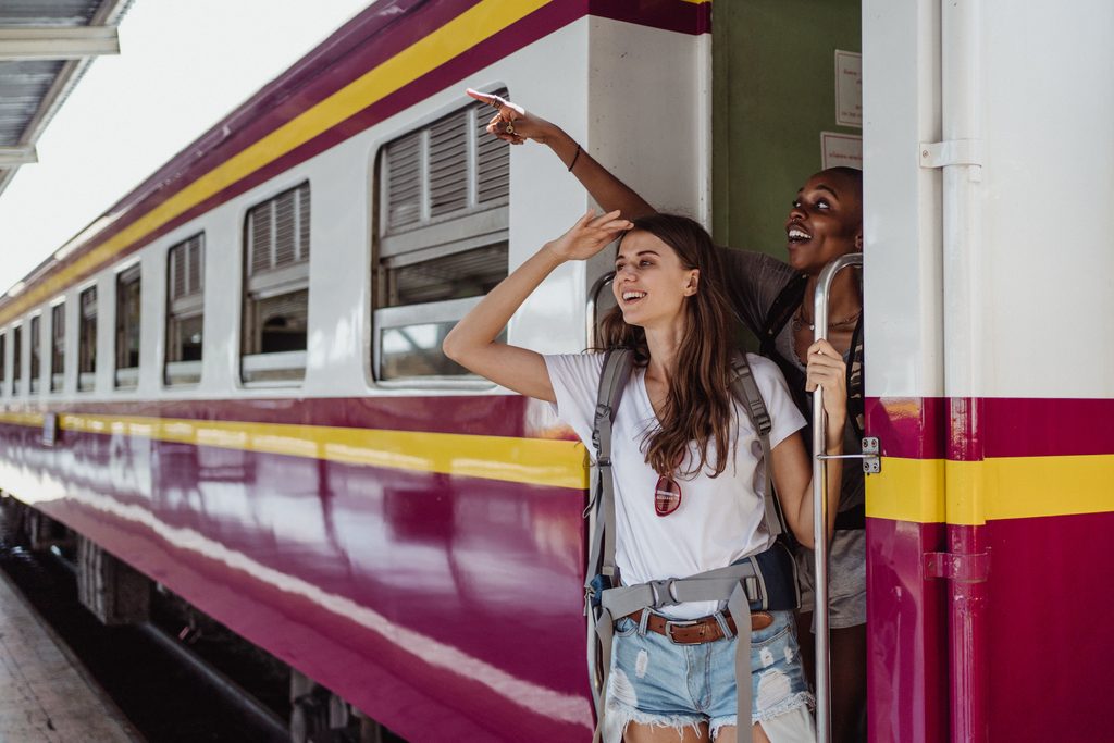 Two smiling travelers with backpacks standing in the doorway of a colorful passenger train before a scenic railway adventure.