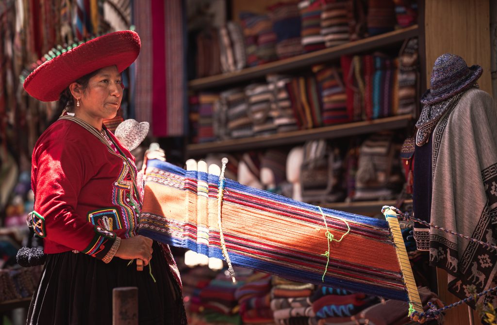 Andean woman wearing traditional red clothing and a wide-brimmed hat beside a loom displaying colorful handwoven textiles in Peru.