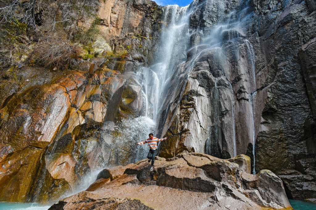 Traveler standing on large rocks beneath a towering waterfall cascading down rugged cliffs into a turquoise pool.