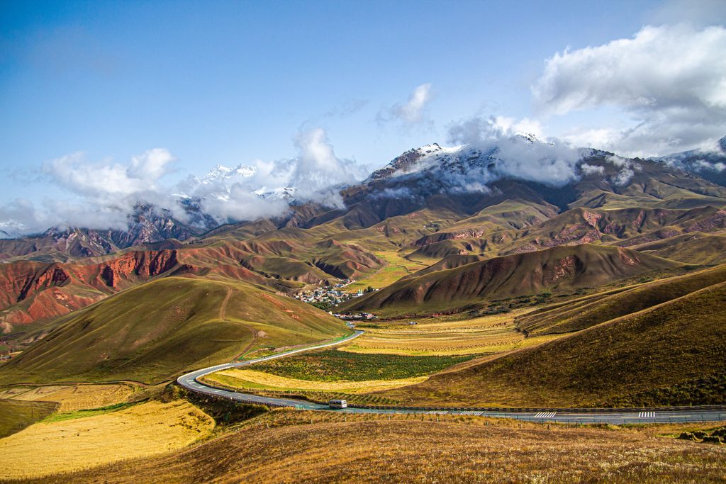 Winding mountain road passing through golden agricultural fields and rolling Andean hills, with snowcapped mountains and clouds in the distance.