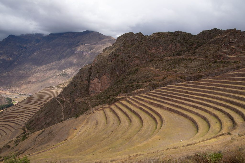 Ancient Inca agricultural terraces stretching across dry golden hillsides in Peru's Sacred Valley, surrounded by rugged Andean mountains beneath cloudy skies.