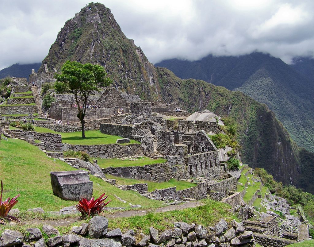 Ancient Inca stone ruins and green terraces at Machu Picchu, Peru, with Huayna Picchu mountain rising above the archaeological site beneath dramatic clouds.
