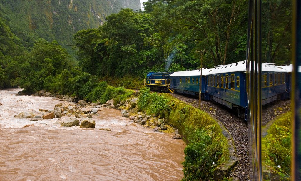 Blue PeruRail train traveling around a mountain curve beside the rushing Urubamba River, surrounded by lush green forests on the scenic railway route to Machu Picchu, Peru.