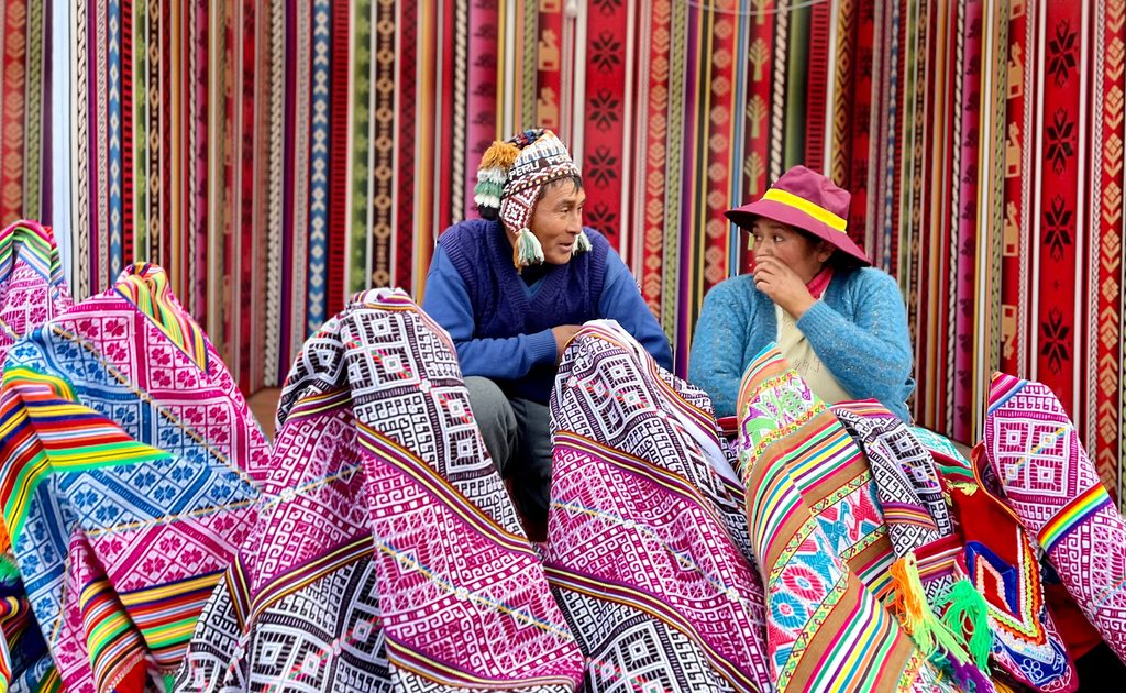 Two Andean textile vendors sitting among colorful handwoven blankets, patterned fabrics, and traditional Peruvian textiles at a market.